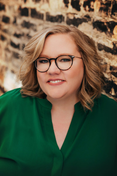 a person with glasses and a green shirt standing in front of a brick wall